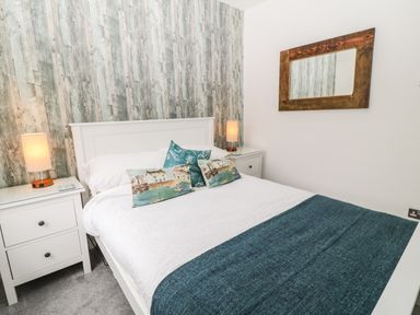 A bedroom with a double bed with white bedding and blue throw pillows between two white nightstands with lamps and a wooden framed mirror on the wall at Harbour Hideaway in Looe
