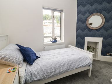 A bedroom with a white bed and blue bedding a window with a sill and a patterned blue feature wall with a round mirror and a small fireplace at Harbour Hideaway in Looe