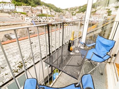 A small balcony with two blue folding chairs and a table with a bottle and two glasses overlooking rooftops and a hillside town at Harbour Hideaway in Looe