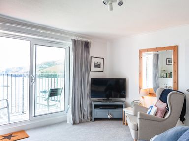 A living room with a large window and sliding door leading to a balcony with a chair a TV on a stand and an armchair next to a side table with a lamp at 32 The Salcombe in Salcombe