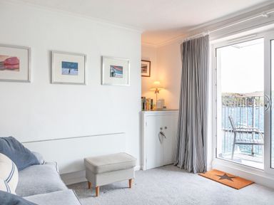 A living room with a sofa ottoman framed pictures and a sliding door opening to a balcony with a chair and a sea view at 32 The Salcombe in Salcombe