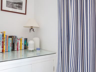 A corner with a white cabinet holding books a lamp and decorative items next to striped blue and white curtains at 32 The Salcombe in Salcombe
