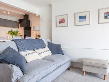 A living room with a gray sofa and cushions next to a small ottoman and kitchen in the background at 32 The Salcombe in Salcombe