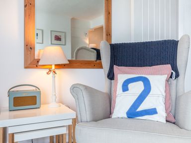 A beige armchair with two cushions and a dark blanket next to a white side table with a lamp and a radio at 32 The Salcombe in Salcombe