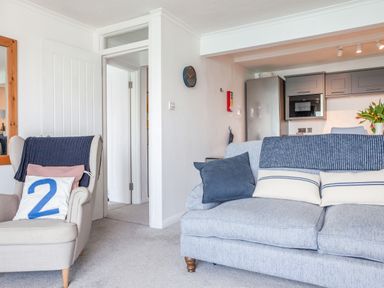A living room with a beige armchair and a grey sofa with cushions near a kitchen at 32 The Salcombe in Salcombe