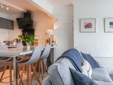 A kitchen area with bar stools and a vase of flowers next to a sofa with cushions and a blanket at 32 The Salcombe in Salcombe