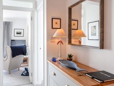 A hallway with a dresser topped with a lamp, mirror, plant, decorative dish, and guestbook with a view into a room with an armchair and television at 32 The Salcombe in Salcombe