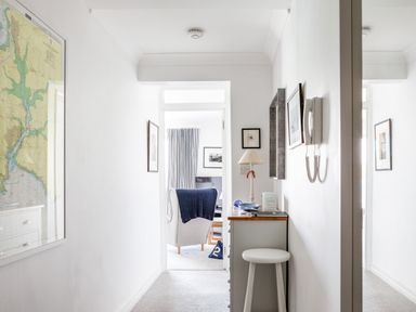 A hallway with framed pictures on the walls a chest of drawers with a lamp and a stool leading to a room with a chair and a television at 32 The Salcombe in Salcombe
