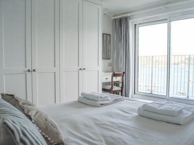 A bedroom with a bed towels on top a wardrobe and a chair by a window overlooking water at 32 The Salcombe in Salcombe