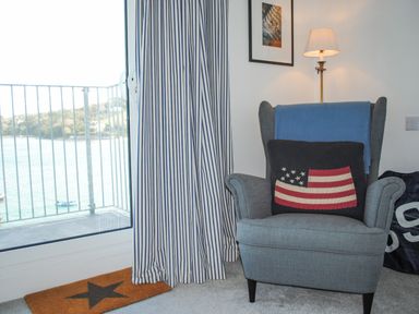 A gray armchair with a pillow featuring an american flag design next to a window with striped curtains and a floor lamp at 32 The Salcombe in Salcombe
