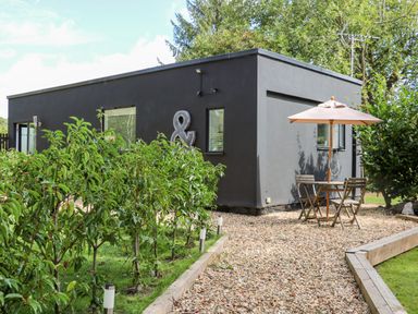 A dark gray modern house with small windows surrounded by greenery and a gravel patio with a wooden table and chairs under a beige umbrella at Kub House in Wigginton Hertfordshire