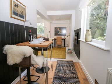 A dining area with a small wooden table set for two and a chair with a white fur throw a second table with a wicker chair is against the far wall with a framed poster above a large window and a vase on the window sill at Kub House in Wigginton Hertfordshire