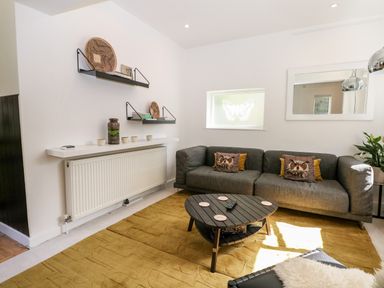 A living room with a gray sofa with owl cushions a triangular coffee table on a yellow rug and wall shelves at Kub House in Wigginton Hertfordshire