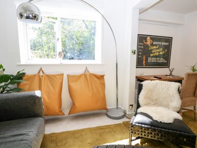 A living room with a gray sofa two orange cushions a large window a tall floor lamp and a black chair with a white fur throw at Kub House in Wigginton Hertfordshire