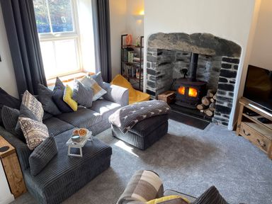 A living room with a gray sofa and cushions a stone fireplace with a wood stove and fire a television and wooden shelf at 7 Castle Terrace in Dolwyddelan