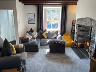 A living room with a sofa and cushions next to a stone fireplace with a fire at 7 Castle Terrace in Dolwyddelan