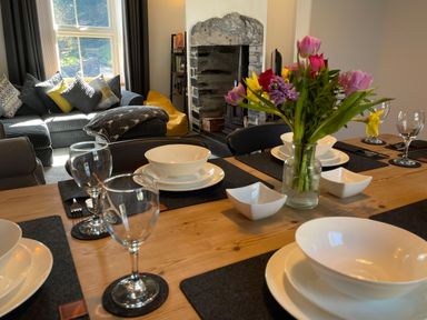 A dining table set with bowls glasses and a flower vase in a living room with a sofa fireplace and window at 7 Castle Terrace in Dolwyddelan