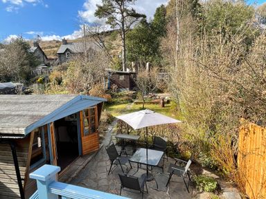 A garden with a patio table and chairs under an umbrella next to a small wooden shed surrounded by trees and bushes at 7 Castle Terrace in Dolwyddelan