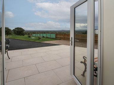 An open glass door leading to a tiled patio with a bench and a view of a lawn and distant landscape at Border View Lodge in Llandrinio