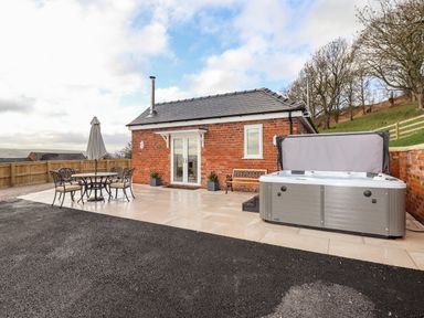 A small brick building with a patio area containing a table with four chairs and an umbrella and a hot tub next to a bench at Border View Lodge in Llandrinio