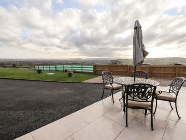 An outdoor patio with a metal table and four chairs with cushions and a closed umbrella next to a paved path and grassy area at Border View Lodge in Llandrinio