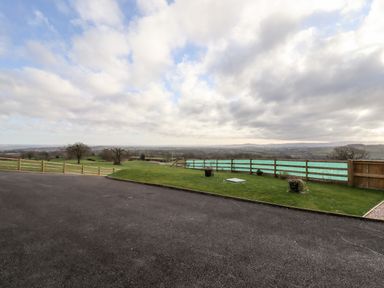 A paved driveway with a fenced grassy area and a distant view of fields and trees at Border View Lodge in Llandrinio