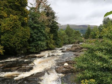 A river with flowing water surrounded by trees and rocks with mountains in the background at Millmore Cabin in Killin
