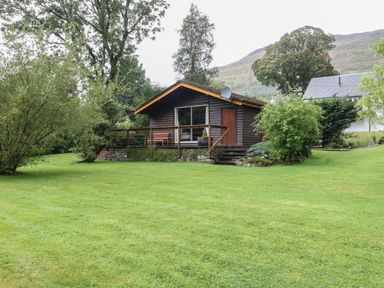 A small wooden cabin with a deck and a bench surrounded by grass and trees at Millmore Cabin in Killin