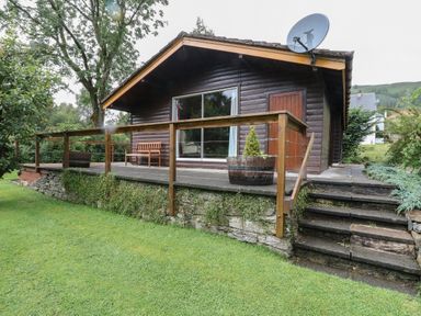 A wooden cabin with a patio area bench and potted plants and stairs leading to a grassy lawn at Millmore Cabin in Killin