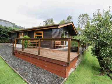 A wooden cabin with a raised deck and railing surrounded by grass and trees at Millmore Cabin in Killin