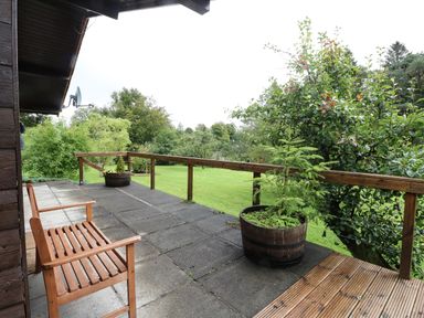 A wooden bench and two planters on a concrete patio with wooden railing and green lawn and trees beyond at Millmore Cabin in Killin