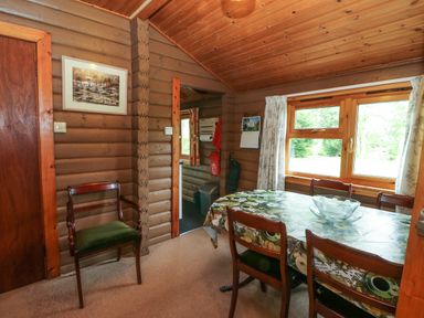 A dining room with wooden walls and ceiling a table covered with a patterned tablecloth and wooden chairs by a window at Millmore Cabin in Killin