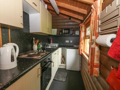 A kitchen with black countertops wooden cabinets a white kettle stove microwave and window with curtains at Millmore Cabin in Killin