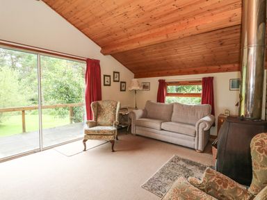 A living room with armchairs a sofa wood stove and large window at Millmore Cabin in Killin