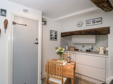 A small kitchen area with a wooden table and chair a bottle of wine and flowers on the table at Cobblers Cottage in Upton Upon Severn