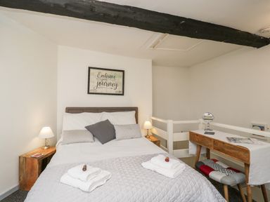 A bedroom with a bed with pillows and folded towels on a quilt a wooden side table with a lamp a wooden desk with a mirror and magazines and a patterned bench at Cobblers Cottage in Upton Upon Severn