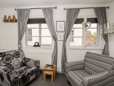 A sitting area with two patterned armchairs a small wooden nested table and two windows with grey curtains at Cobblers Cottage in Upton Upon Severn