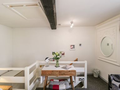A small wooden desk with flowers and a mirror in a white room with stairs and a patterned stool at Cobblers Cottage in Upton Upon Severn