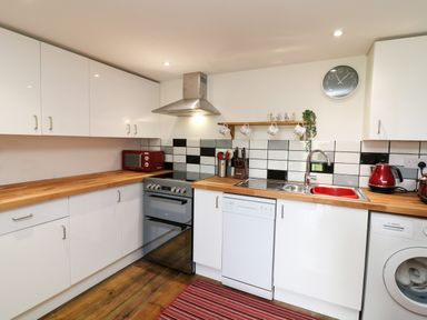 A kitchen with white cabinets wooden countertops a silver oven a red microwave a sink with a red basin a washing machine and utensils on a striped rug at Wisteria Cottage in Marldon
