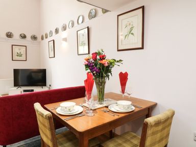 A dining table with two place settings and a vase of flowers alongside a red sofa and a television on a white stand at Wisteria Cottage in Marldon