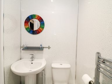A small bathroom with white sink and toilet a colorful round mirror and a glass shelf with toiletry items at Wisteria Cottage in Marldon