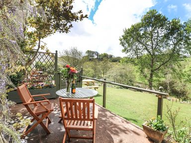 An outdoor patio with wooden chairs and a table with flowers and a bottle overlooking a grassy garden with trees at Wisteria Cottage in Marldon