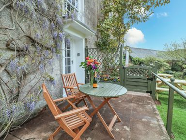A small patio with wooden chairs and a round table with flowers and a bottle outside a stone wall with wisteria at Wisteria Cottage in Marldon