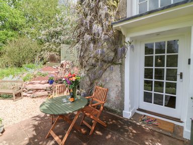 A patio with a round table, two wooden chairs, a bench, flowers, and a glass door at Wisteria Cottage in Marldon