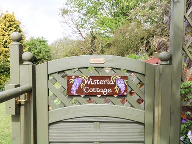 A green wooden garden gate with a sign saying Wisteria Cottage and plants in the background at Wisteria Cottage in Marldon