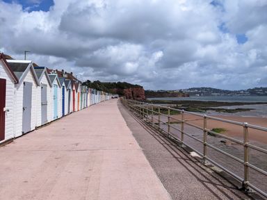 A row of colorful beach huts along a paved walkway next to a metal railing and a rocky beach with water and cliffs in the distance