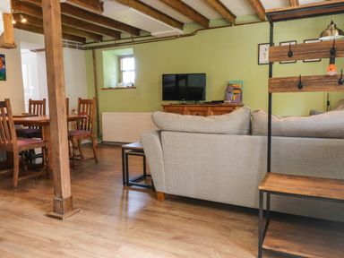 A living room with a gray sofa a wooden dining table with chairs a television on a wooden cabinet and exposed wooden beams on the ceiling at Bwthyn Gwyn Ty'n Y Pant in Boduan near Nefyn