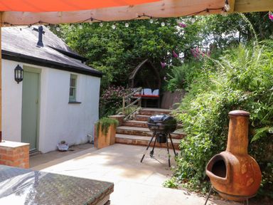 An outdoor patio area with a small white building a barbecue grill stairs with a wooden seating area and greenery at Bwthyn Gwyn Ty'n Y Pant in Boduan near Nefyn