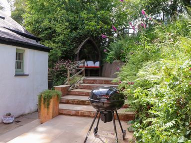 An outdoor patio area with a barbecue grill steps leading to a wooden bench surrounded by greenery at Bwthyn Gwyn Ty'n Y Pant Boduan near Nefyn