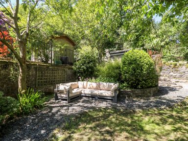 An outdoor garden area with a sectional sofa on a stone pathway surrounded by trees and bushes at Yr Hen Ysgol The Old School in Prenteg